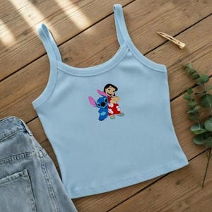 May include: Light blue ribbed tank top featuring a graphic of Lilo and Stitch. The cartoon characters are hugging each other. The top has thin straps and a scoop neckline. A pair of denim shorts is visible on the left side of the image.
