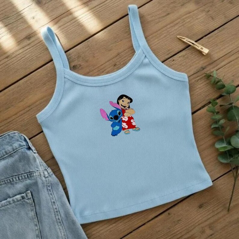 May include: Light blue ribbed tank top featuring a graphic of Lilo and Stitch. The cartoon characters are hugging each other. The top has thin straps and a scoop neckline. A pair of denim shorts is visible on the left side of the image.