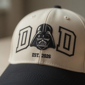 Puede incluir: Una gorra de béisbol beige con visera negra que presenta un texto bordado que dice DAD, con un icono negro del casco de Darth Vader reemplazando la letra A. Debajo del icono, el texto EST. 2026 aparece bordado en negro en el panel frontal.