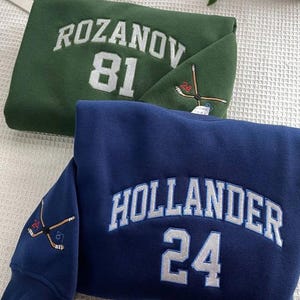 May include: Two sweatshirts, one green and one navy blue, are folded. The green sweatshirt has "ROZANOV 81" embroidered on the front. The navy blue sweatshirt has "HOLLANDER 24" embroidered on the front. Both have hockey stick details.