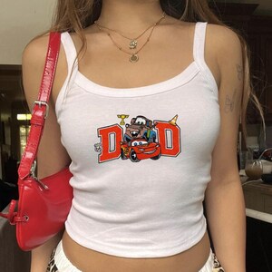 K&ouml;nnte beinhalten: Wei&szlig;es Tanktop mit einem Cars-Motiv. Das Motiv zeigt das Wort "DAD" in roten Blockbuchstaben, mit Zeichentrickautos und einem Route 66-Schild. Eine rote Umh&auml;ngetasche ist ebenfalls zu sehen.
