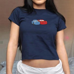 May include: Navy blue crop top featuring an embroidered design of two cartoon cars, one blue and one red. The short-sleeved shirt is made of a soft material and has a round neckline. The cars are smiling and appear to be from the movie "Cars".