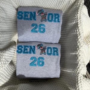 May include: Two gray sweatshirts with the text "SENIOR 26" in teal glitter lettering and a cartoon character. The sweatshirts are folded and stacked on top of each other. The background is a white knitted blanket.