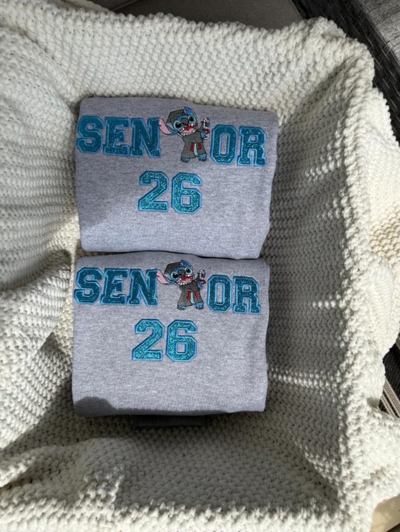 May include: Two gray sweatshirts with the text "SENIOR 26" in teal glitter lettering and a cartoon character. The sweatshirts are folded and stacked on top of each other. The background is a white knitted blanket.