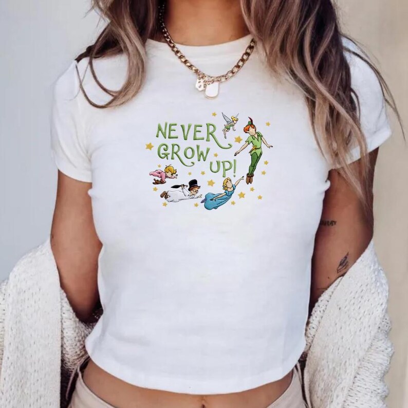 Peut inclure: Un t-shirt blanc &agrave; manches courtes avec une impression graphique des personnages de Peter Pan et le texte "NEVER GROW UP!" en lettres vertes, entour&eacute; de petites &eacute;toiles jaunes. Le motif inclut la F&eacute;e Clochette et Peter Pan.