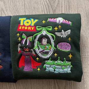 May include: Dark green sweatshirt featuring embroidered characters and the "Toy Story" logo. The design includes Buzz Lightyear, aliens, and other characters from the animated film. The sweatshirt has a crew neck and long sleeves.