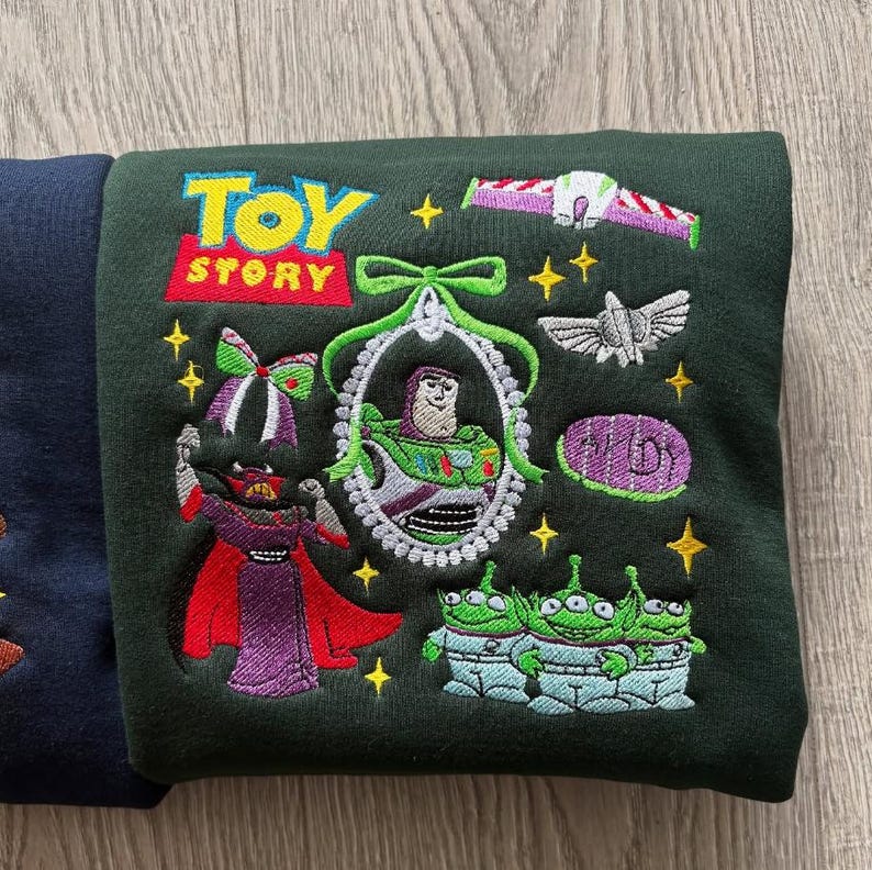 May include: Dark green sweatshirt featuring embroidered characters and the "Toy Story" logo. The design includes Buzz Lightyear, aliens, and other characters from the animated film. The sweatshirt has a crew neck and long sleeves.