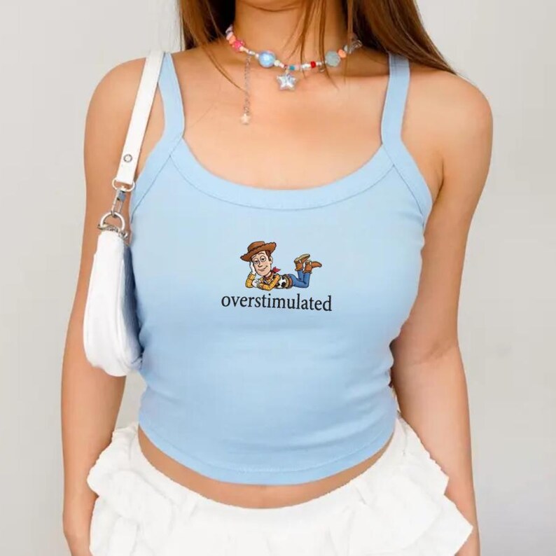 K&ouml;nnte beinhalten: Hellblaues Tanktop mit Spaghettitr&auml;gern. Das Shirt zeigt eine Cartoon-Grafik von Woody aus Toy Story und das Wort "overstimulated" in schwarzer Schrift. Ein l&auml;ssiger Stil.