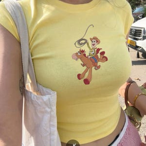 May include: A yellow, short-sleeved t-shirt featuring a cartoon graphic of Woody and Bullseye from Toy Story. The graphic shows Woody riding Bullseye with a lasso. The shirt is a fitted style.