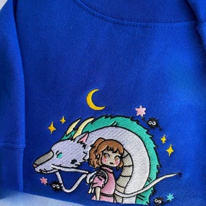 May include: Royal blue sweatshirt featuring an embroidered design of a dragon, a girl, stars, and a crescent moon. The design is in shades of white, green, pink, yellow, and black. The sweatshirt has a crew neck and long sleeves.