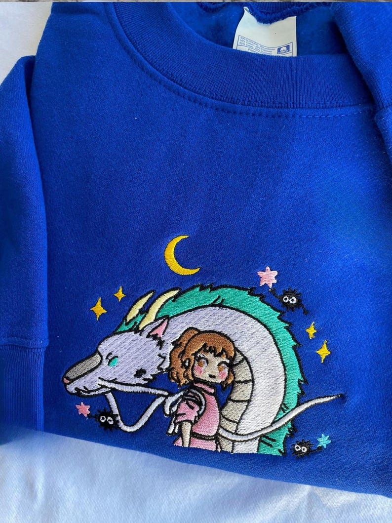May include: Royal blue sweatshirt featuring an embroidered design of a dragon, a girl, stars, and a crescent moon. The design is in shades of white, green, pink, yellow, and black. The sweatshirt has a crew neck and long sleeves.