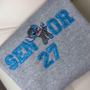 May include: A gray sweatshirt with the word "SENIOR" and the number "27" in blue glitter lettering. A cartoon character is also featured. The sweatshirt is folded.