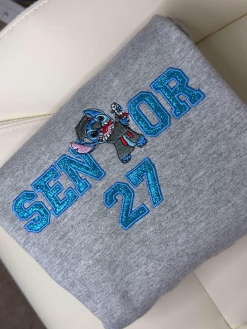 May include: A gray sweatshirt with the word "SENIOR" and the number "27" in blue glitter lettering. A cartoon character is also featured. The sweatshirt is folded.