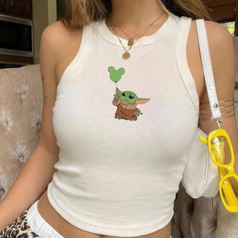 May include: A white sleeveless ribbed vest top featuring a central graphic of a small green creature with large ears wearing a brown robe and holding a green balloon shaped like a mouse head. The top has a high crew neckline and a fitted silhouette.