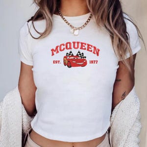 Puede incluir: Camiseta blanca con un gr&aacute;fico rojo de un coche de dibujos animados y el texto "MCQUEEN EST. 1977". El dise&ntilde;o incluye banderas a cuadros. La camiseta es de cuello redondo.