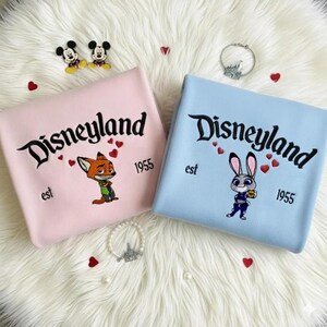Op de afbeelding: Twee sweatshirts, &eacute;&eacute;n roze en &eacute;&eacute;n blauw, met "Disneyland" geborduurd in zwart schrift. Het roze sweatshirt heeft een vos-personage en de blauwe een konijn-personage. Beide hebben "est 1955" eronder.