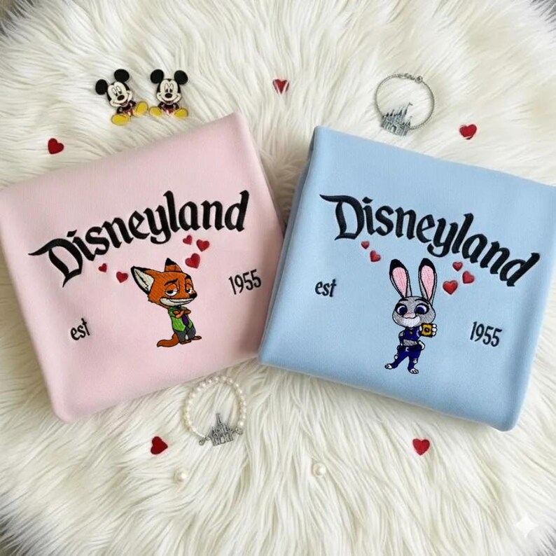 Op de afbeelding: Twee sweatshirts, &eacute;&eacute;n roze en &eacute;&eacute;n blauw, met "Disneyland" geborduurd in zwart schrift. Het roze sweatshirt heeft een vos-personage en de blauwe een konijn-personage. Beide hebben "est 1955" eronder.