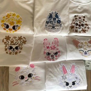 May include: White t-shirts with embroidered animal faces. Designs include a chick, a bunny, a bear, and a fox, each decorated with floral accents in various colors. The embroidery adds a handcrafted touch to the shirts.