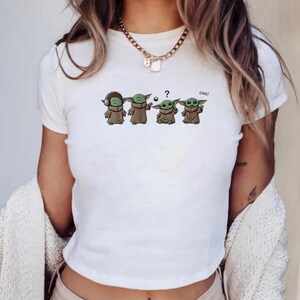 May include: White t-shirt featuring a cartoon graphic of Grogu in various poses. The Grogu characters are wearing headphones, holding a ball, and reading a book. The word "coo!" is printed above one of the characters.