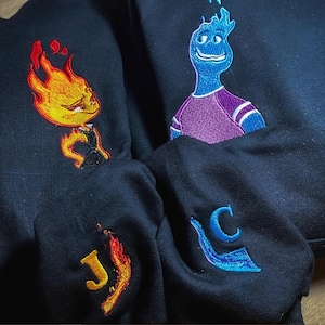 Può includere: Felpe nere con personaggi dei cartoni animati ricamati. Una presenta una figura infuocata, l'altra un personaggio blu simile all'acqua. Le maniche mostrano iniziali ricamate. Le felpe sono di colore scuro.