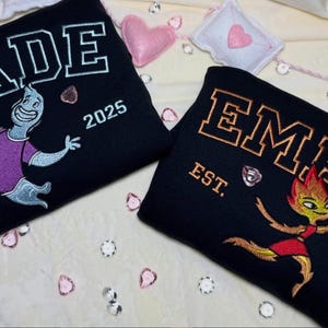 Puede incluir: Dos sudaderas negras con diseños bordados. Una dice "WADE" con un personaje de dibujos animados y "2025". La otra dice "EMBER" con un personaje de dibujos animados y "2025". Ambas tienen "EST." debajo del nombre.