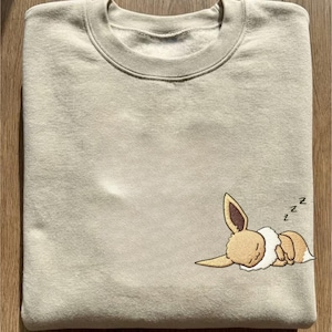 Op de afbeelding: Een beige sweatshirt met een ronde hals. Het heeft een geborduurd ontwerp van een slapend stripfiguur met bruine en witte vacht. Het ontwerp bevat de letter "z" om slaap aan te duiden.