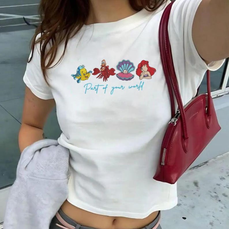 May include: White t-shirt featuring a colorful graphic of characters from a popular animated film. The design includes a yellow fish, a red crab, a pink seashell, and a red-haired mermaid, with the text "Part of your world" in light blue.