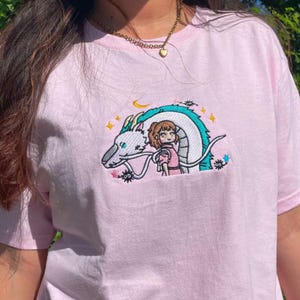 May include: A light pink t-shirt featuring an embroidered design of a white dragon with a turquoise mane and a girl in a pink dress. The design includes stars and a crescent moon. The shirt is made of soft material.