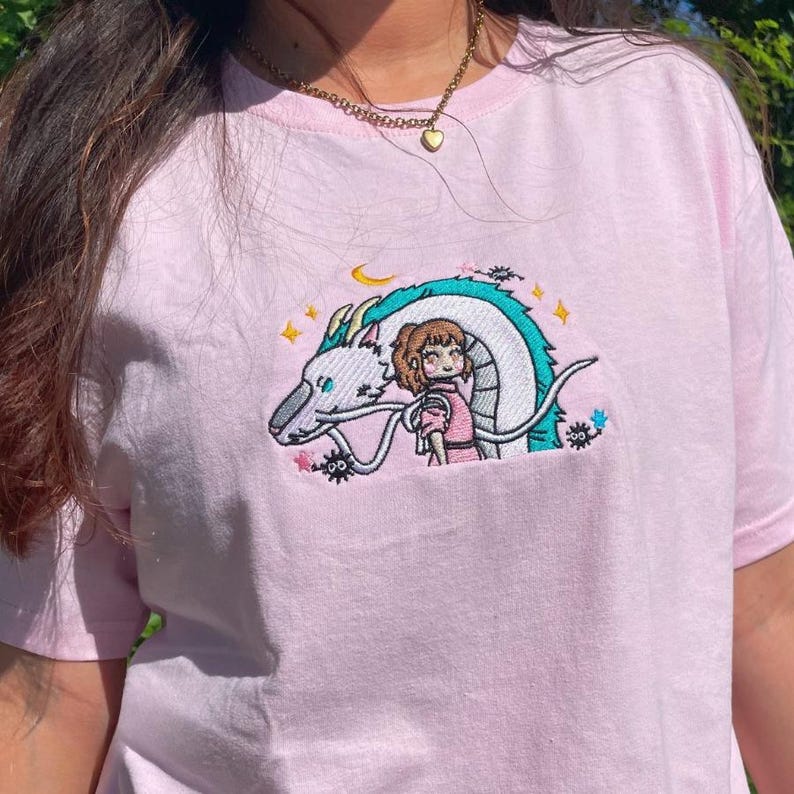 May include: A light pink t-shirt featuring an embroidered design of a white dragon with a turquoise mane and a girl in a pink dress. The design includes stars and a crescent moon. The shirt is made of soft material.