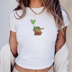 May include: A white short-sleeved t-shirt featuring a small embroidered graphic of a green creature with large ears wearing a brown robe and holding a green balloon shaped like a mouse head. The shirt has a crew neckline and a simple fitted cut.
