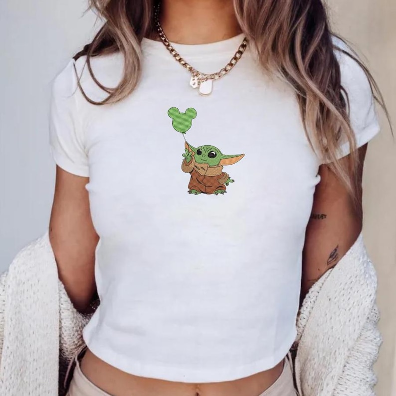May include: A white short-sleeved t-shirt featuring a small embroidered graphic of a green creature with large ears wearing a brown robe and holding a green balloon shaped like a mouse head. The shirt has a crew neckline and a simple fitted cut.