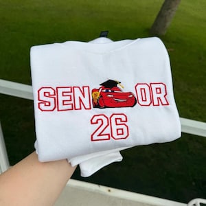 Puede incluir: Sudadera blanca con texto bordado en rojo y un dibujo animado de un coche. El texto dice "SENIOR 26". El dibujo del coche es rojo con un birrete. Un gran regalo para un graduado de secundaria.