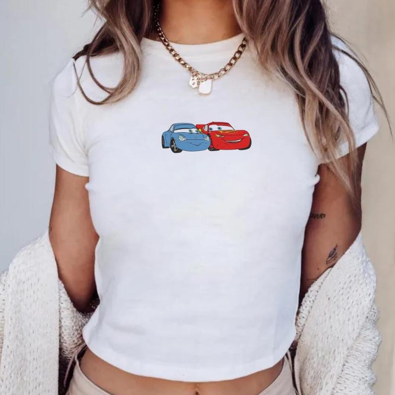 May include: White t-shirt featuring a cartoon graphic of two cars, one blue and one red. The cars are side-by-side, with the red car on the right. The shirt is a crew neck style.