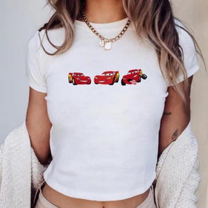 Puede incluir: Camiseta blanca con un dise&ntilde;o de tres coches de carreras rojos de dibujos animados. Los coches est&aacute;n en diferentes poses, uno de ellos sacando la lengua. La camiseta es de estilo corto.