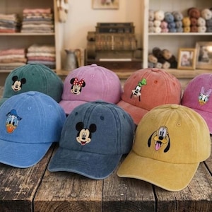 Op de afbeelding: Een verzameling van zeven kleurrijke baseballpetten met Disney-figuren. De petten zijn in verschillende kleuren, waaronder blauw, groen, roze en geel, elk geborduurd met een ander personage zoals Mickey Mouse en Donald Duck.
