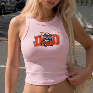 K&ouml;nnte beinhalten: Hellrosa geripptes Tanktop mit einem Cartoon-Auto-Motiv und dem Wort "DAD" in orangefarbenen Blockbuchstaben. Das Design umfasst Route 66-Schilder und eine Troph&auml;e. Das Top ist &auml;rmellos.
