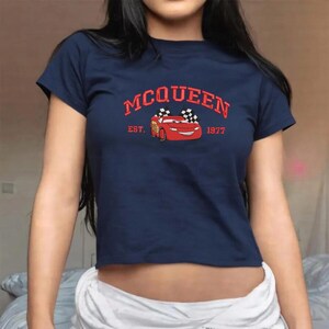 Puede incluir: Camiseta azul marino con un gr&aacute;fico de coche de dibujos animados rojo. El texto "MCQUEEN" est&aacute; arriba y "EST. 1977" abajo. El coche tiene banderas a cuadros. Cuello redondo, mangas cortas.