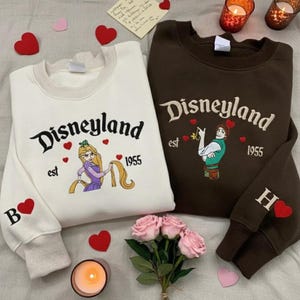 Könnte beinhalten: Zwei Sweatshirts, eines in Creme und eines in Braun, mit "Disneyland" auf der Vorderseite gestickt. Das cremefarbene Sweatshirt zeigt Rapunzel, das braune zeigt Flynn Rider. Beide haben "est 1955" und Herzdetails. Das cremefarbene Sweatshirt hat ein "B" und ein Herz auf dem Ärmel, das braune ein "H".