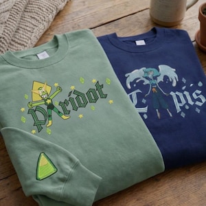 Può includere: Due felpe girocollo, una verde salvia e una blu navy, con disegni di personaggi dei cartoni animati. La felpa verde presenta la parola "Peridot" e una grafica del personaggio. La felpa blu ha una grafica del personaggio e la parola "Lapis".