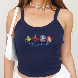 May include: Navy blue tank top with a scoop neck and thin straps. The front features a colorful graphic of characters from The Little Mermaid, with the text "Part of your world" in a script font.
