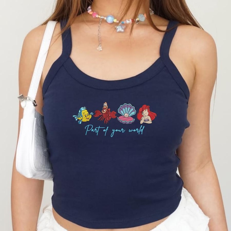 May include: Navy blue tank top with a scoop neck and thin straps. The front features a colorful graphic of characters from The Little Mermaid, with the text "Part of your world" in a script font.