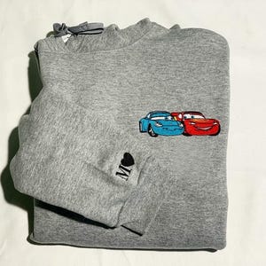 May include: A grey sweatshirt with an embroidered design of two cartoon cars, one blue and one red. The sleeve has a small black heart with the letter "M" embroidered on it. The sweatshirt is folded neatly.