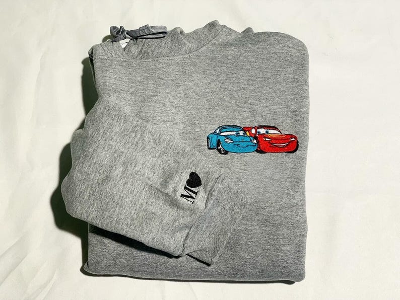 May include: A grey sweatshirt with an embroidered design of two cartoon cars, one blue and one red. The sleeve has a small black heart with the letter "M" embroidered on it. The sweatshirt is folded neatly.