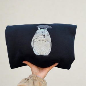 May include: A black sweatshirt featuring an embroidered image of Totoro, a character from the animated film My Neighbor Totoro. The design shows Totoro holding an umbrella. The sweatshirt is held by a person.