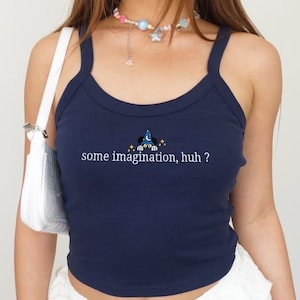 May include: Navy blue tank top with a scoop neck and spaghetti straps. The shirt features the text "some imagination, huh?" with a small graphic of a wizard hat and Mickey Mouse ears. The top is worn with a white purse.
