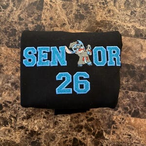 May include: Black fabric item with the text "SENIOR 26" in light blue glitter lettering. A cartoon character is embroidered between the words "SENIOR" and "26". The item is folded and appears to be a piece of clothing.
