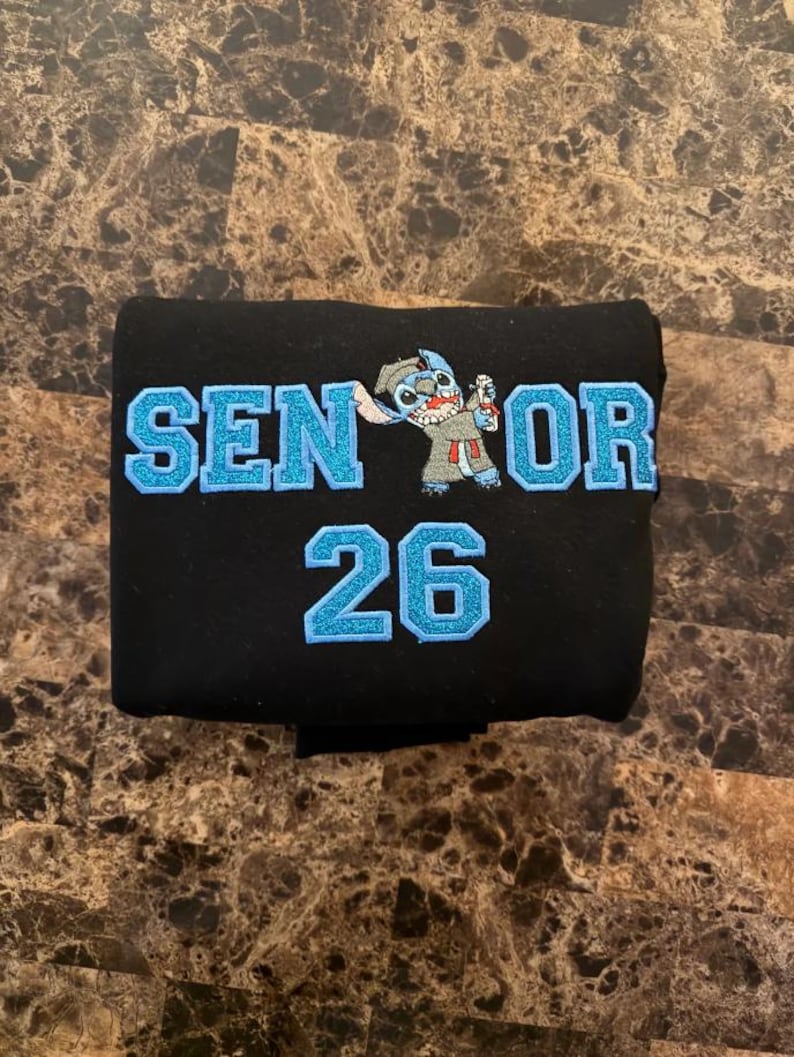 May include: Black fabric item with the text "SENIOR 26" in light blue glitter lettering. A cartoon character is embroidered between the words "SENIOR" and "26". The item is folded and appears to be a piece of clothing.