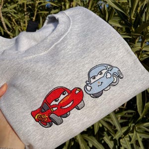 Puede incluir: Sudadera gris claro con coches de dibujos animados bordados. Un coche es rojo con el n&uacute;mero 95, y el otro es azul claro. La sudadera est&aacute; hecha de un material suave y es adecuada para uso casual.