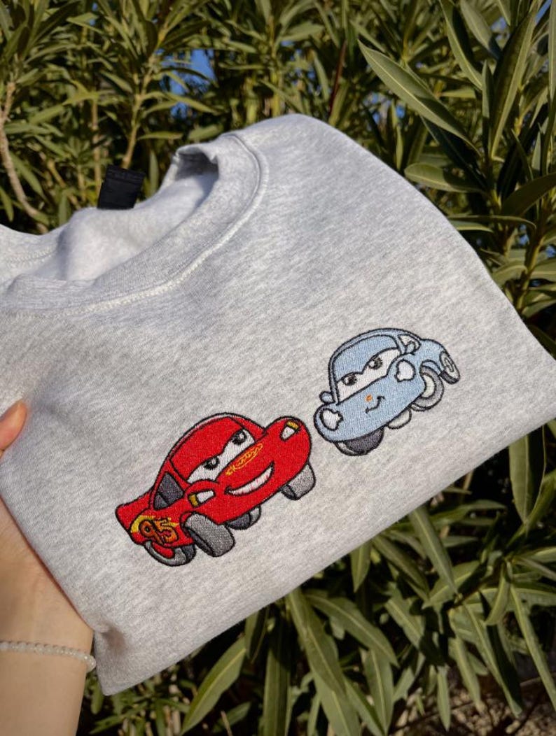 Puede incluir: Sudadera gris claro con coches de dibujos animados bordados. Un coche es rojo con el n&uacute;mero 95, y el otro es azul claro. La sudadera est&aacute; hecha de un material suave y es adecuada para uso casual.