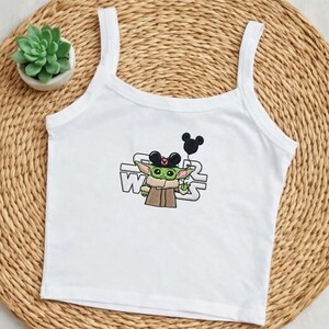 May include: White tank top with a graphic of Baby Yoda wearing Mickey Mouse ears and holding a black balloon. The Star Wars logo is behind the character. A small succulent plant in a white pot sits to the left.